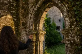 Detail of ancient stone arches with moss, warm sunlight, and a solitary figure admiring medieval architecture in a quiet courtyard