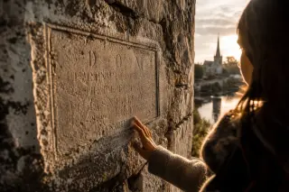 Weathered stone wall with historic inscriptions highlighted by soft golden light, a visitor gently touching the surface in quiet admiration