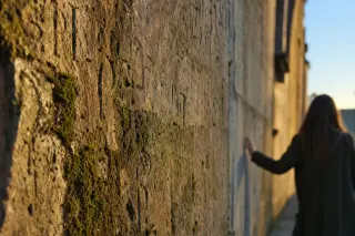 Historic stone wall in Wassy with old engravings, softly lit by late afternoon sun, a person seen from behind gently touching the weathered surface in quiet reflection.