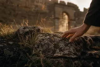 Moss-covered historic stones half-buried in grass, gently touched by a hand near a faded old gate in warm late afternoon light