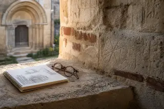 Faded stone wall with old engravings and a sketchbook with glasses on a window ledge, gentle afternoon light, arched doorway softly blurred in the background.