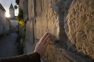 Hand tracing artisan marks on an aged limestone wall in a quiet, golden-lit village alleyway