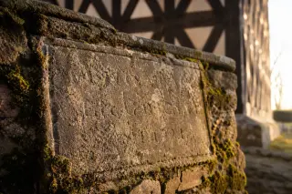 Textured stone plaque with moss at the base of an old timber belfry, softly lit by late afternoon sun, evoking quiet contemplation of local heritage.